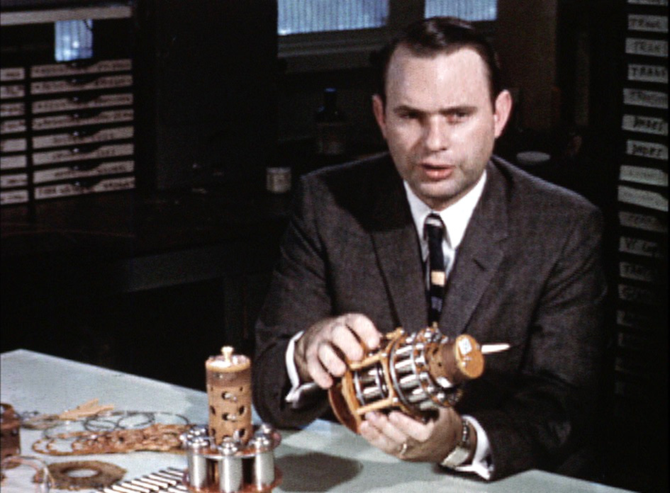 Henry Richter, in a dark suit, explains a cylindrical satellite instrument package at a JPL workbench in 1957